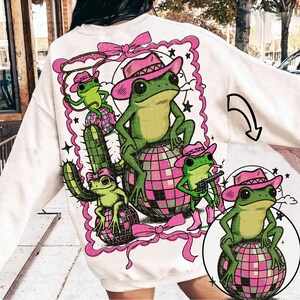 May include: White sweatshirt featuring a whimsical design of green frogs wearing pink cowboy hats, boots, and holding a lasso and a gun. The frogs are sitting on disco balls and a cactus, surrounded by a pink frame and bows.