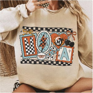 Op de afbeelding: Beige sweatshirt met een kleurrijk grafisch ontwerp. Het ontwerp bevat de letters "M" en "A" in postzegelachtige kaders, een bliksemschicht, dobbelstenen, kersen en een vlinder. Het ontwerp is omlijnd met een zwart-wit geblokt patroon.