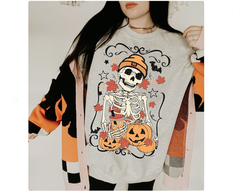 May include: A gray sweatshirt with a skeleton wearing a hat and holding a pumpkin. The skeleton is surrounded by pumpkins and fall leaves. The text "Shroom Queens" is printed on the sweatshirt.