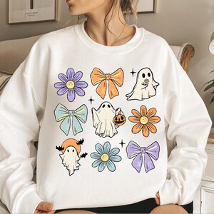 May include: White sweatshirt with a colorful Halloween design featuring ghosts, bows, and flowers. The design includes a ghost holding a pumpkin, a ghost with a flower, and a ghost with bats.