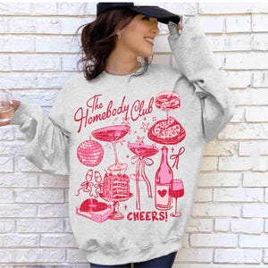 May include: A gray sweatshirt with a pink graphic design that says "The Homebody Club" and features a wine bottle, glasses, pizza, and a disco ball. The text "Cheers!" is also included in the design.