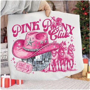 May include: White pillow with a pink graphic design featuring a cowboy hat, disco ball, and horses. The text "PINK PONY CLUB" is displayed above the hat. An "ADMIT ONE" ticket is also visible.