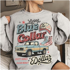 May include: A light gray sweatshirt with a graphic design featuring a blue pickup truck, cowboy boots, and a baseball cap. The text reads "Living Blue Collar Dollar" and "Dirty Hands Clean Money."