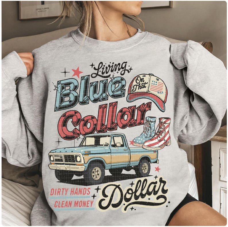 May include: A light gray sweatshirt with a graphic design featuring a blue pickup truck, cowboy boots, and a baseball cap. The text reads "Living Blue Collar Dollar" and "Dirty Hands Clean Money."