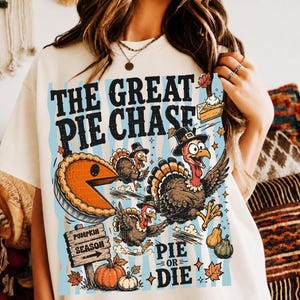 May include: A cream-colored t-shirt featuring a graphic with the text THE GREAT PIE CHASE and PIE OR DIE. The design includes illustrations of turkeys wearing hats, a large orange pumpkin pie, a wooden sign labeled PUMPKIN SEASON, and pumpkins.