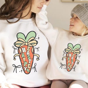 May include: White sweatshirts with a carrot graphic and the text "Happy Easter" written vertically on the carrot. The carrot is green at the top and has a yellow bow.