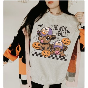 Puede incluir: Una sudadera gris con un gr&aacute;fico de dos vacas de dibujos animados que llevan sombreros y sostienen calabazas. El texto "Howdy Fall" est&aacute; impreso en la sudadera.