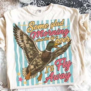 May include: Cream-colored t-shirt featuring a graphic of a duck in flight with the text "Some glad Morning when this life is over I'll Fly Away." The design includes blue, pink, yellow, and brown colors. The shirt is a Comfort Colors brand.