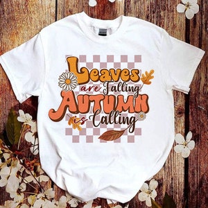 Op de afbeelding: Wit T-shirt met een grafische print van oranje en bruine tekst die "Leaves are falling Autumn is calling" luidt, met een geruite achtergrond en bloemmotieven.