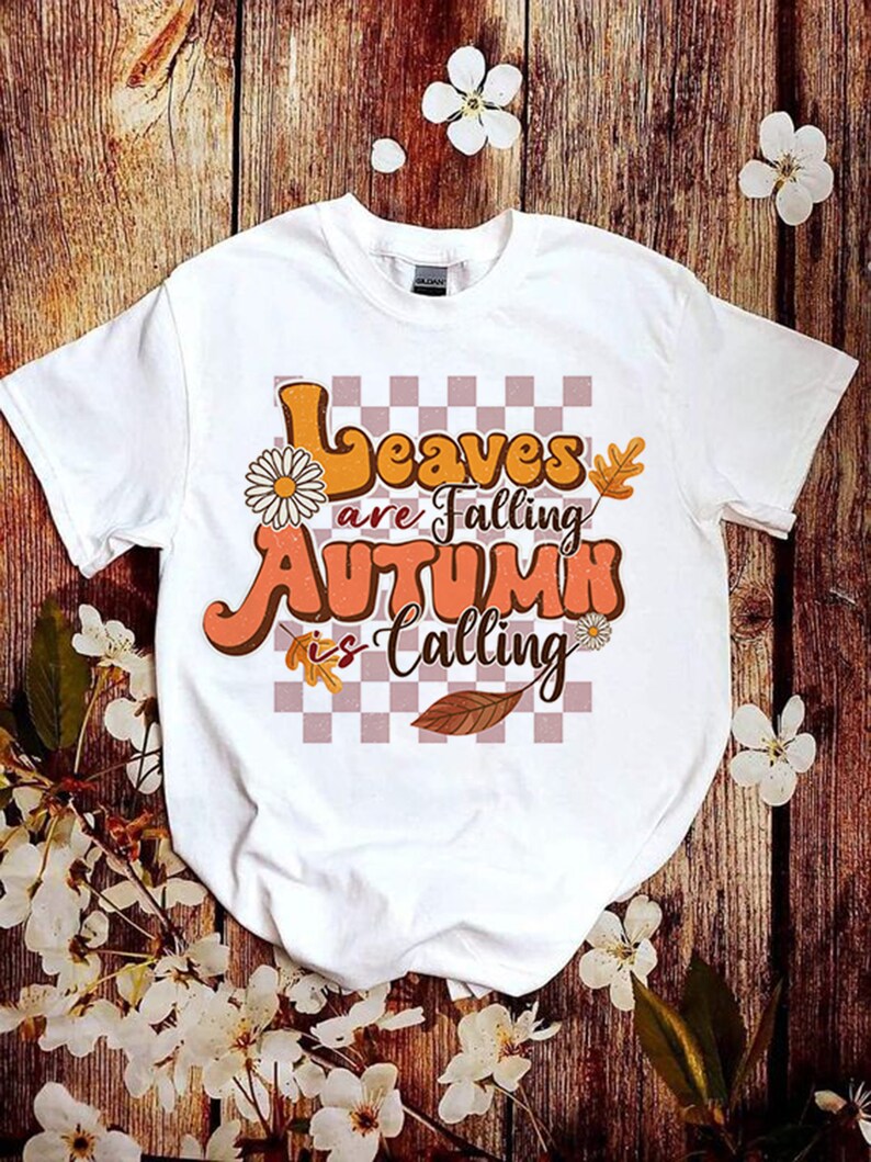 Op de afbeelding: Wit T-shirt met een grafische print van oranje en bruine tekst die "Leaves are falling Autumn is calling" luidt, met een geruite achtergrond en bloemmotieven.