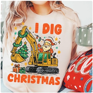 May include: A light beige sweatshirt with the text "I DIG CHRISTMAS" in orange. The graphic features Santa Claus driving a yellow excavator decorated with a Christmas tree and lights. Presents are at the bottom.