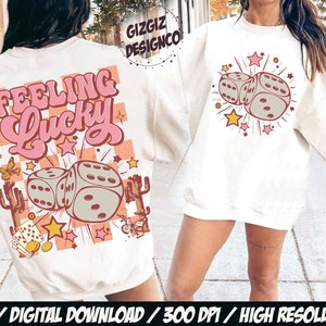 May include: Cream-colored sweatshirt with a graphic design featuring two dice, stars, and the text "Feeling Lucky" in pink and peach. The design includes cacti and butterflies. The sweatshirt is a digital download.