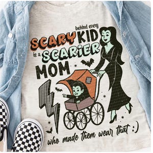 May include: A light gray t-shirt with a Halloween-themed graphic. The design features the text "Scary Kid is a Scarier Mom" with a cartoon illustration of a vampire baby in a stroller being pushed by a woman. The text "who made them wear that :)" is also included.