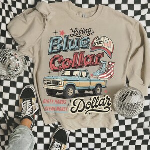 May include: Beige crewneck sweatshirt with a graphic design featuring a blue and yellow pickup truck, cowboy boots, and the text "Living Blue Collar Dollar." The design has a vintage, retro aesthetic.
