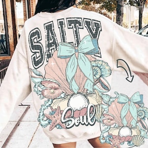 Puede incluir: Una sudadera de color crema con un estampado gr&aacute;fico de una concha con una perla, una estrella de mar y un lazo. El texto "Salty Soul" est&aacute; impreso en la sudadera.