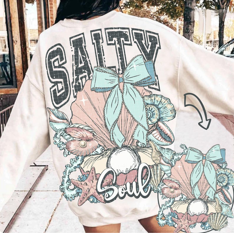Puede incluir: Una sudadera de color crema con un estampado gr&aacute;fico de una concha con una perla, una estrella de mar y un lazo. El texto "Salty Soul" est&aacute; impreso en la sudadera.
