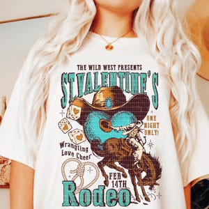 以下が含まれることがあります： ヴィンテージスタイルのグラフィックデザインが施された白いTシャツ。カウボーイが馬に乗っている様子、サイコロ、そして「The Wild West Presents St. Valentine's One Night Only! Feb 14th Rodeo」というテキストが描かれています