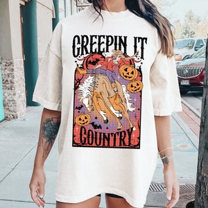 Puede incluir: Camiseta blanca con un gr&aacute;fico de una bruja montando un caballo con calabazas y murci&eacute;lagos. El texto "Creepin' It Country" est&aacute; impreso en la camiseta.