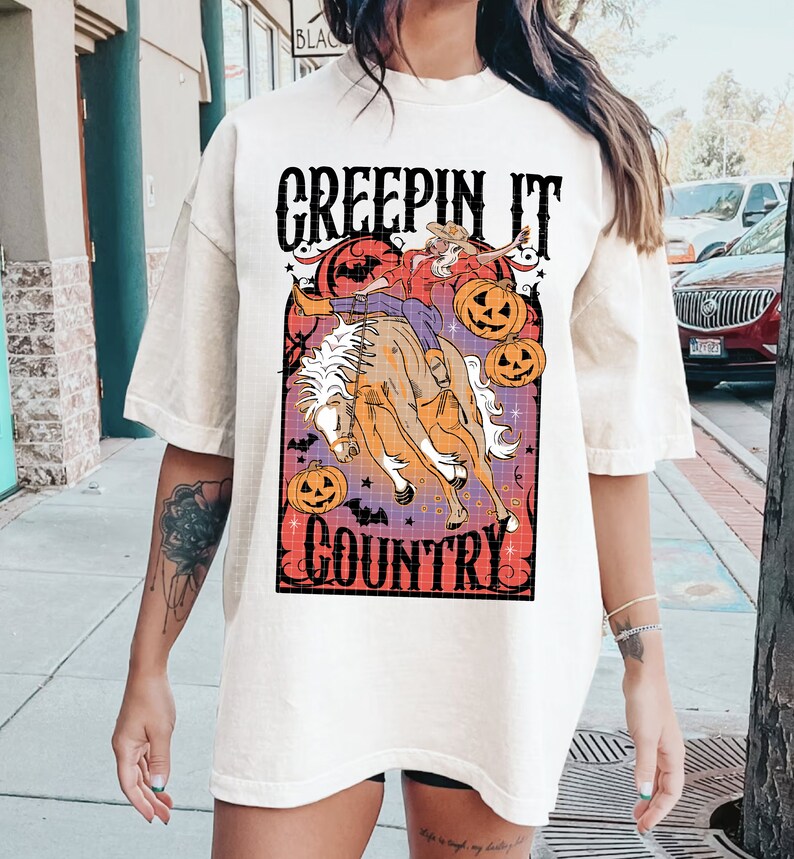 Puede incluir: Camiseta blanca con un gr&aacute;fico de una bruja montando un caballo con calabazas y murci&eacute;lagos. El texto "Creepin' It Country" est&aacute; impreso en la camiseta.