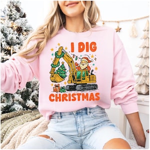 May include: A light pink sweatshirt with the text "I DIG CHRISTMAS" in orange. The graphic features Santa Claus operating a yellow excavator decorated with a Christmas tree and lights. Presents are at the bottom.