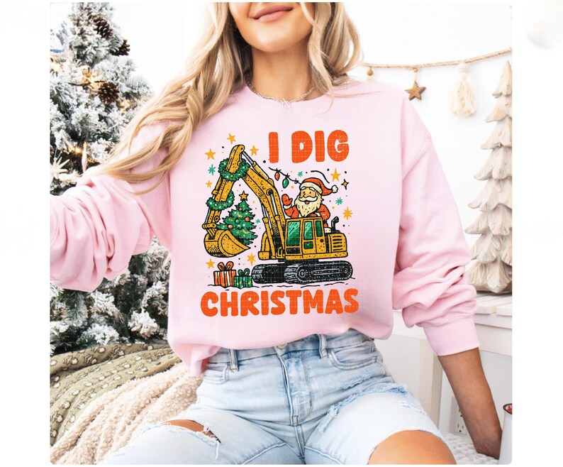 May include: A light pink sweatshirt with the text "I DIG CHRISTMAS" in orange. The graphic features Santa Claus operating a yellow excavator decorated with a Christmas tree and lights. Presents are at the bottom.