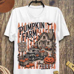 Peut inclure: Un t-shirt blanc avec une impression graphique repr&eacute;sentant une ferme de citrouilles. Le motif inclut un vieux camion, une maison en pierre, des citrouilles, des chauves-souris et un croissant de lune. Le texte indique PUMPKIN FARM, PICK YOUR PUMPKIN FRESHLY GROWN et MAGICALLY GROWN.