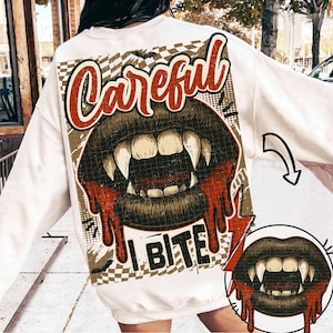 May include: White sweatshirt with a graphic of a vampire mouth with fangs, dripping blood, and the words "Careful I Bite". The design has a vintage, distressed look with a checkered background.