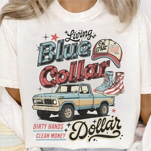 May include: White t-shirt with a graphic design featuring a blue and red vintage truck, boots, and a baseball cap. The text reads "Living Blue Collar Dollar" with additional text "Dirty Hands Clean Money".