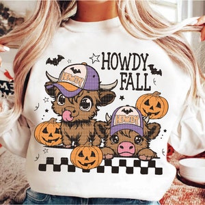 Puede incluir: Sudadera blanca con un gr&aacute;fico de dos vacas Highland de dibujos animados que llevan sombreros de vaquero y sostienen calabazas. El texto "Howdy Fall" est&aacute; impreso sobre las vacas.