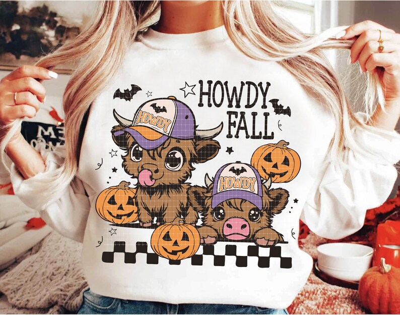 Puede incluir: Sudadera blanca con un gr&aacute;fico de dos vacas Highland de dibujos animados que llevan sombreros de vaquero y sostienen calabazas. El texto "Howdy Fall" est&aacute; impreso sobre las vacas.