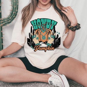 May include: A white t-shirt with a graphic design of a pumpkin with turquoise jewels and the text "Howdy Pumpkin".