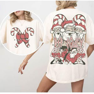 May include: A cream-colored t-shirt with a Christmas-themed graphic on the front. The graphic features three Santa Clauses with candy canes and the text "Da Jolly". The back of the shirt features a red bow with candy canes.