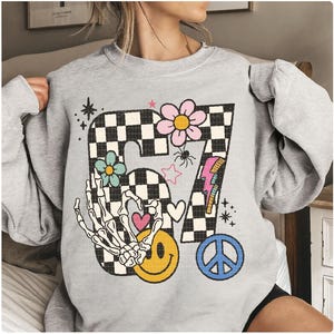 May include: A light gray sweatshirt with a large graphic featuring the number 67 in a checkered pattern. The design includes a skeleton hand, flowers, a smiley face, a peace sign, and a lightning bolt. The sweatshirt has long sleeves.