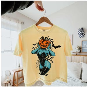 May include: Yellow short-sleeve t-shirt featuring a Halloween-themed graphic. The design includes a jack-o'-lantern head, a black corset, and blue skin. The shirt is hanging on a wooden hanger.