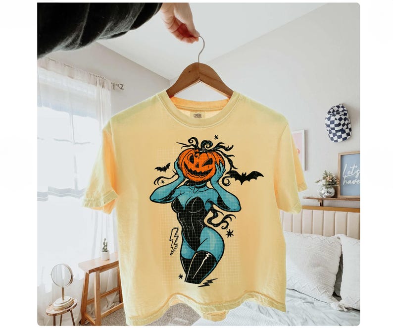 May include: Yellow short-sleeve t-shirt featuring a Halloween-themed graphic. The design includes a jack-o'-lantern head, a black corset, and blue skin. The shirt is hanging on a wooden hanger.