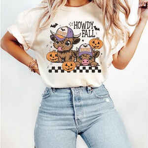 Puede incluir: Una camiseta blanca con un gr&aacute;fico de dos vacas de dibujos animados que llevan sombreros y sostienen calabazas. El texto "Howdy Fall" est&aacute; impreso en la camiseta.