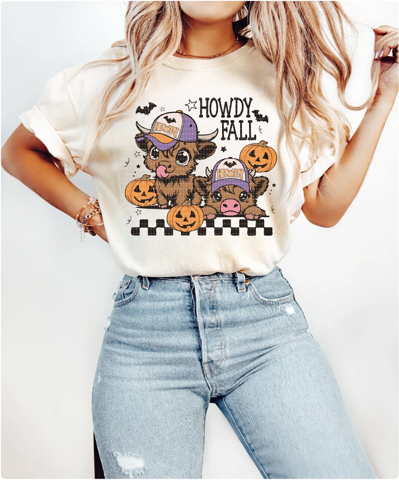 Puede incluir: Una camiseta blanca con un gr&aacute;fico de dos vacas de dibujos animados que llevan sombreros y sostienen calabazas. El texto "Howdy Fall" est&aacute; impreso en la camiseta.