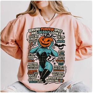 May include: A light pink sweatshirt with a graphic featuring a pumpkin-headed witch with text that reads "Serving Pumpkin Spice and Deadly Looks." The design includes phrases like "Cute but Creepy" and "Wicked Never Looked This Good."