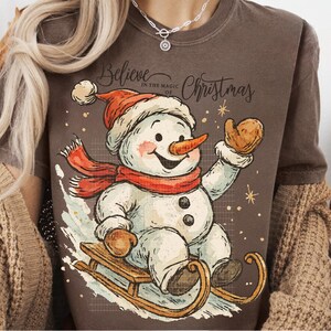 Op de afbeelding: Bruin T-shirt met een vrolijke sneeuwman met een rode sjaal en hoed, op een houten slee. De sneeuwman heeft een wortelneus en is omringd door sneeuwvlokken. De tekst "Believe in the magic of Christmas" staat erboven.