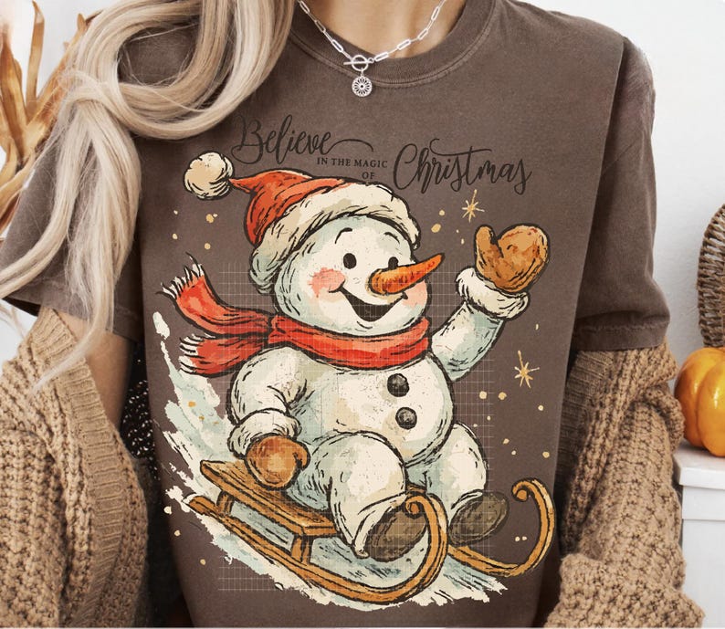 Op de afbeelding: Bruin T-shirt met een vrolijke sneeuwman met een rode sjaal en hoed, op een houten slee. De sneeuwman heeft een wortelneus en is omringd door sneeuwvlokken. De tekst "Believe in the magic of Christmas" staat erboven.