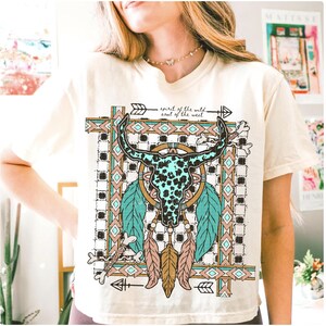 以下が含まれることがあります： クリーム色のTシャツ。 ターコイズと黒の雄牛の頭蓋骨のデザイン、羽、矢印が描かれています。 デザインの上には「spirit of the wild, soul of the west」の文字がプリントされています。 シャツは半袖でゆったりとしたフィット感です。