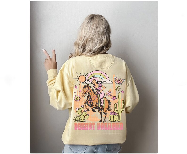 Pu&ograve; includere: Una felpa gialla con una stampa grafica di una donna a cavallo in un paesaggio desertico. La grafica include un arcobaleno, un sole, cactus e stelle. Il testo "DESERT DREAMER" &egrave; stampato sotto la grafica.