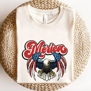 May include: A white t-shirt with a red, white, and blue American flag design featuring an eagle with the word "Merica" in a retro font.