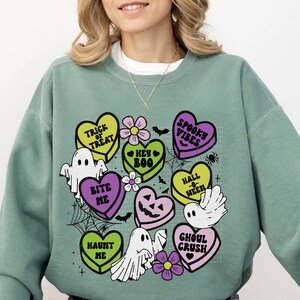 May include: Green sweatshirt with a graphic print of candy hearts with Halloween-themed sayings like "Trick or Treat", "Bite Me", and "Spooky Vibes".