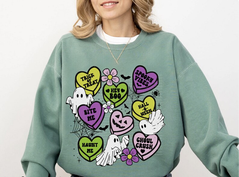 May include: Green sweatshirt with a graphic print of candy hearts with Halloween-themed sayings like "Trick or Treat", "Bite Me", and "Spooky Vibes".