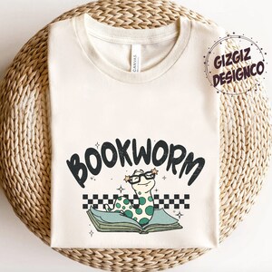 Pu&ograve; includere: Una maglietta bianca con un verme verde che indossa gli occhiali e legge un libro. Il testo "BOOKWORM" &egrave; sopra il verme. La maglietta &egrave; su una superficie intrecciata.