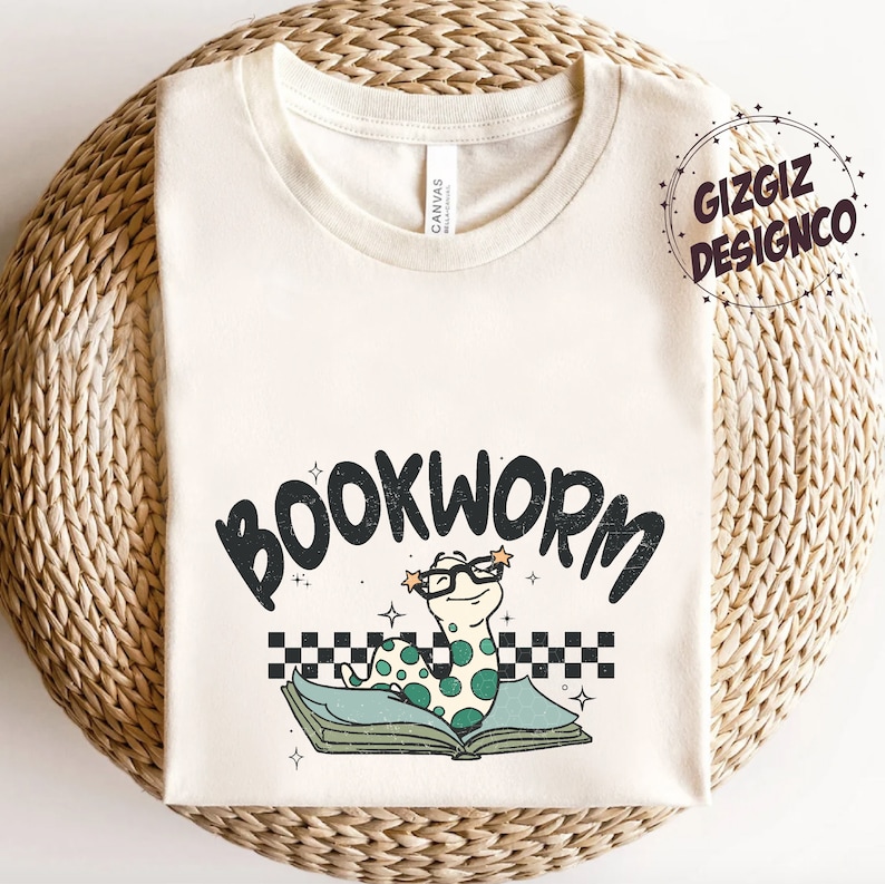 Pu&ograve; includere: Una maglietta bianca con un verme verde che indossa gli occhiali e legge un libro. Il testo "BOOKWORM" &egrave; sopra il verme. La maglietta &egrave; su una superficie intrecciata.