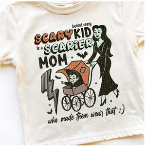 May include: Cream-colored t-shirt with a Halloween-themed graphic. The design features a cartoon vampire child in a stroller pushed by a woman in a black dress. Text reads "Behind every scary kid is a scarier mom who made them wear that :) "