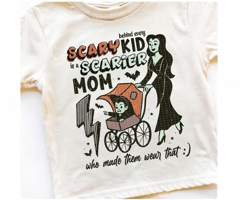 May include: Cream-colored t-shirt with a Halloween-themed graphic. The design features a cartoon vampire child in a stroller pushed by a woman in a black dress. Text reads "Behind every scary kid is a scarier mom who made them wear that :) "