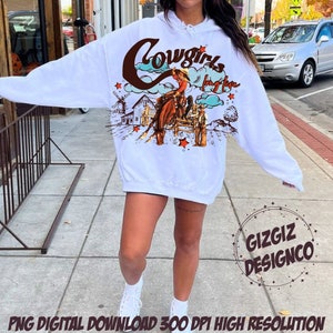 May include: White hoodie with a western-themed graphic. The design features a cowgirl riding a horse, the words "Cowgirls Long Live", and a rural landscape with a windmill and barn. The hoodie also has the text "GIZGIZ DESIGNCO".